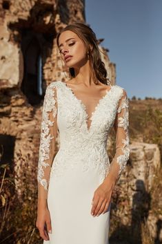 a woman wearing a wedding dress with sheer sleeves and an open back, standing in front of ruins