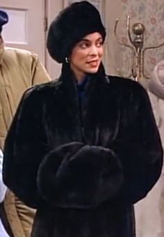 a woman in a black fur coat standing next to another man wearing a brown hat