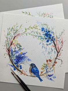 watercolor painting of two blue birds in a floral wreath on white paper next to a fountain pen