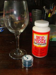 a wine glass next to a bottle of modd podge on a wooden table