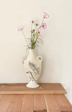 a white vase with pink flowers in it sitting on a wooden table next to a wall