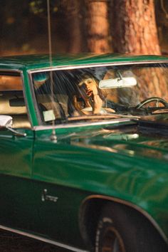 a dog sitting in the driver's seat of a green car with trees in the background