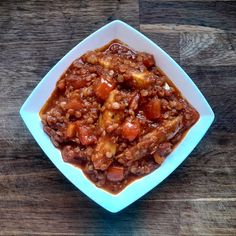 a white bowl filled with chili and meat on top of a wooden table next to a fork