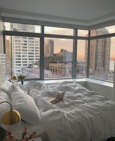 a bed with white sheets and pillows in a bedroom next to large windows overlooking the city