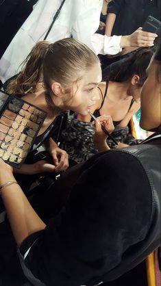 two women sitting at a table with makeup brushes in their mouths and one woman brushing her teeth