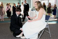 a man kneeling down next to a woman in a white dress on a chair with other people