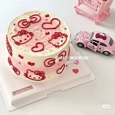 a hello kitty birthday cake with pink car next to it on a white tablecloth