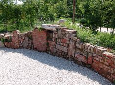 a stone wall made out of bricks in the middle of a gravel area with trees and bushes behind it