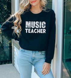 The Music Teacher Sweatshirt is the perfect unisex garment for any music teacher, whether they teach piano, guitar, choir, or any other instrument. Made from high-quality, soft and comfortable fabric, this sweatshirt is perfect for keeping warm during chilly days in the classroom or at home. Featuring the title "Music Teacher" on the front, this sweatshirt is a great way for any music teacher to show off their passion for music education. It also makes a wonderful end of year gift for music teac Music Teacher Tshirt Ideas, Music-themed Cotton Letter Print Sweatshirt, Music-themed Letter Print Cotton Sweatshirt, Music Teacher Shirt, Music-themed Cotton Sweatshirt With Letter Print, Relaxed Fit Music-themed T-shirt With Crew Neck, Music-themed Relaxed Fit Crew Neck T-shirt, Music-themed Cotton Crew Neck Shirt, Shirt Knot