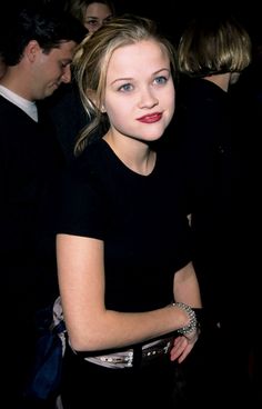 a woman in a black dress standing next to other people and looking at the camera