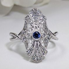 Art Deco Diamond & Blue Sapphire Fashion Ring, Filigree Design 14k White Gold DIAMOND DETAILS: Shape:  Round Cut Diamonds Number of Stones: 44 Carats:  0.20 ct. Clarity: VS-SI - Eye Clean  Color: H-I GEM STONES: Shape:  Round Cut Blue Sapphire Number of stones: 1 Approx. 0.05 ct. Clarity: Eye Clean  Color: Medium Blue SETTING: Material: 14k White Gold (Available Also in 14K Yellow Gold or 14K Pink Gold - No Extra Charge) Ring size 7 (custom made in other sizes) Comes packaged In Pop up gift box Art Deco Blue Diamond Ring With Accents, Art Deco Sapphire Jewelry With Center Stone, Blue Art Deco Diamond Ring With Accents, Heirloom Blue Diamond Ring With Accents, Blue Diamond Ring, Art Deco Style, Gift, Art Deco 14k White Gold Jewelry With Round Cut, Art Deco Diamond White Jewelry With Gemstone, Vintage Diamond White Sapphire Jewelry, Art Deco Diamond White Gemstone Jewelry