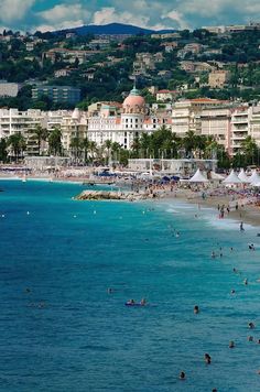 many people are on the beach and in the water near some buildings with hills in the background