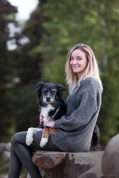 a woman sitting on top of a log holding a dog in her lap and smiling at the camera