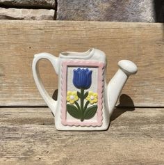 a ceramic teapot with a blue flower painted on the front and side, sitting on a wooden surface