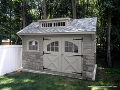 an outdoor storage shed with two doors and windows on the roof, next to a white fence