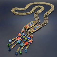 "Millefiori bead fringe necklace of four chains join with a jeweled front clasp set with colorful millefiori art glass tiles. The fringe of brilliant beads are blue, scarlet, aqua, orange, greens and yellow are echoed in the crystal border surrounding the tiles. Bronze metal finish augments the richness of the colors. Millefiori means 'thousand flowers.' Bundles of tiny glass filament rods are sliced, then fired to meld their brilliant colors and floral patterns. No two beads are ever identical. Bead Fringe, Necklace Colorful, Rainbow Jewelry, Hippie Necklace, Glass Tiles, Fringe Necklace, Sweet Romance, Art Deco Necklace, Geometric Necklace