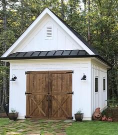 a small white shed with two wooden doors and windows on the roof, in front of some trees