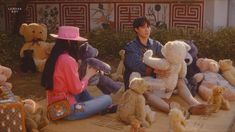 two people sitting on the ground surrounded by stuffed animals and holding teddy bears in their hands