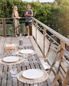 a man and woman standing on a deck next to a table set with plates, glasses and napkins