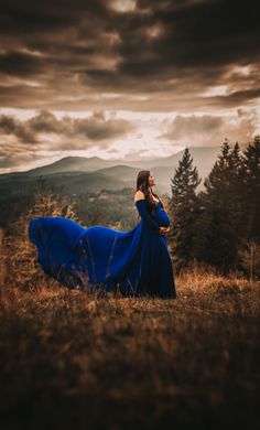 a woman in a blue dress standing on top of a grass covered field under a cloudy sky