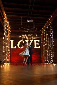 two people standing in front of a love sign with lights on the wall behind them