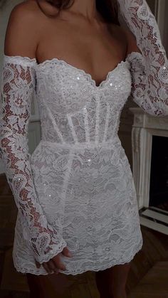 a woman wearing a white dress with long sleeves and sheer lace on the shoulders, standing in front of a fireplace