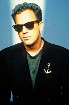 a man wearing sunglasses and a jacket with an anchor pin on his lapel, standing in front of a blue wall