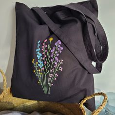 a black tote bag with flowers painted on the front and side, sitting next to a wicker basket