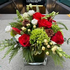 A festive arrangement of seasonal red, white and green flowers, pinecones and berries with lush accenting winter greenery, featured in a clear glass leaf lined cylinder or square vase. White And Green Flowers, Grave Flowers, Winter Greenery