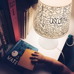 a child's hand on a book next to a lamp and bookshelf