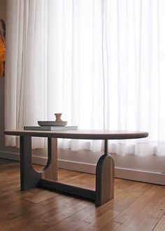 a wooden table sitting in front of a window with white curtains behind it and a bowl on top of the table