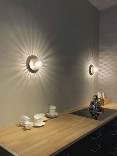 two lights are on the wall above a kitchen counter with cups and saucers in front of it