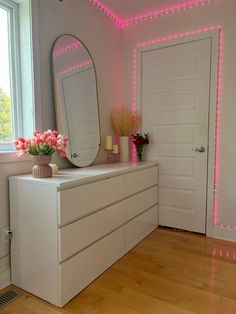 a white dresser sitting next to a large mirror on top of a wooden floor in front of a window