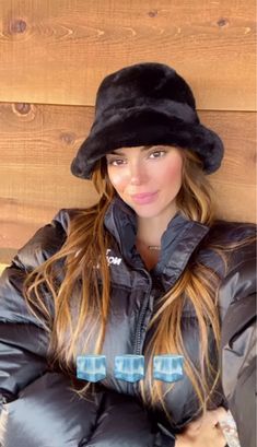 a woman with long hair wearing a black coat and hat, sitting against a wooden wall