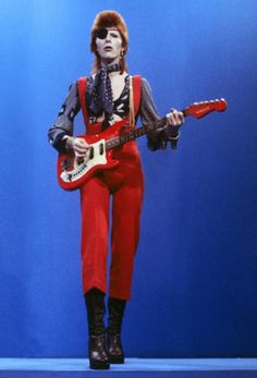 a man in red pants and black boots holding a guitar while standing next to a blue background