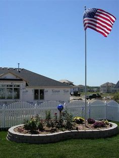 an american flag is flying in front of a white picket fence and flowerbed garden