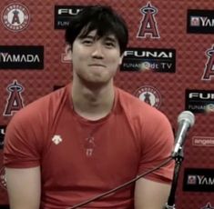 a man sitting at a microphone in front of a red wall with the los angeles angels logo on it