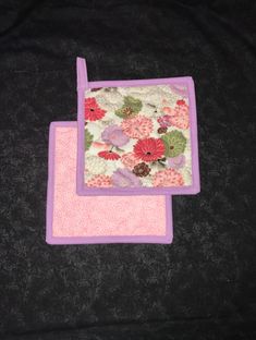 two pot holders with flowers on them sitting on a black cloth covered tablecloth, one is pink and the other is purple