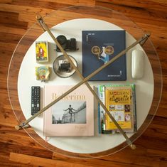 a glass table topped with books and other items on top of a wooden floor next to a white wall