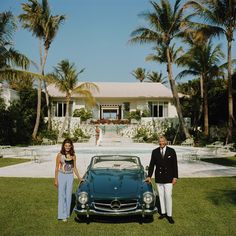 a man and woman standing next to a car in front of a house with palm trees