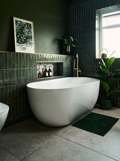 a large white bathtub sitting next to a toilet in a green tiled bathroom with potted plants on the wall