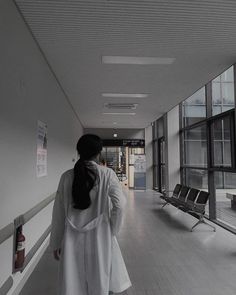 a woman in white coat walking down a long hallway next to a bench and windows