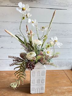 a vase filled with flowers and greenery on top of a wooden table next to a wall