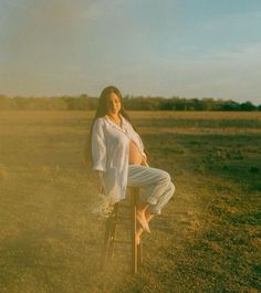 a pregnant woman sitting on top of a wooden stool in an open field at sunset