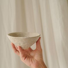 a hand holding a small white bowl in front of a white curtained wall behind it