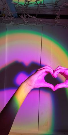 a person's hand making a heart shape in front of a rainbow painted wall