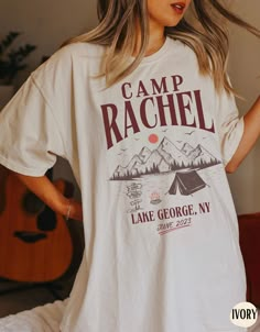 a woman wearing a camp rachel t - shirt standing in front of a bed with her hands on her hips