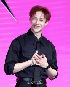 a man in black shirt and tie standing next to a pink wall with his hands together