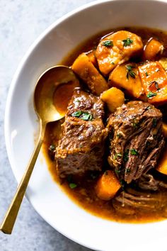 a white bowl filled with meat and potatoes on top of a table next to a spoon