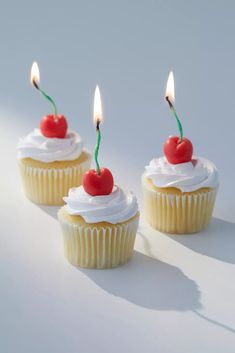 three cupcakes with white frosting and two cherries on top one has a single candle